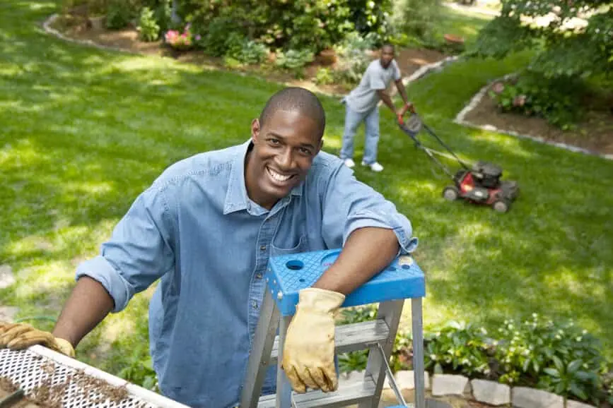A man standing on a ladder smiling as he cleans his gutters.