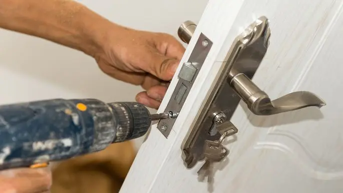 A hand holding a doorknob while replacing the locking mechanism with a drill.