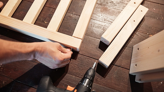 A person uses a power drill to assemble wooden furniture pieces on a wooden surface.