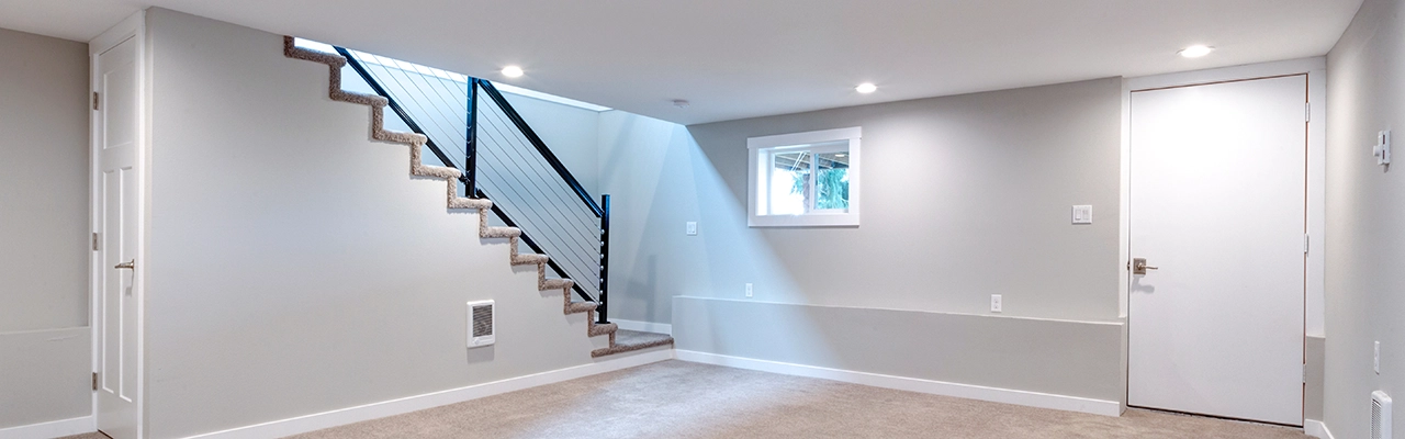 A bright, modern basement with beige carpet, a staircase, a window, and a white door.