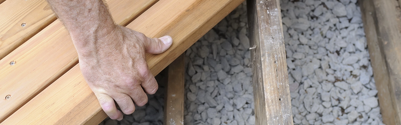 A close-up of a hand placing a wooden plank on a framework of timber slats above gravel.