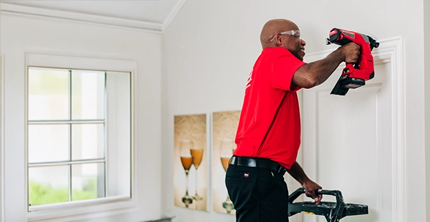 A person in a red shirt uses a nail gun while standing on a ladder to work on a white wall in a bright, modern interior space.