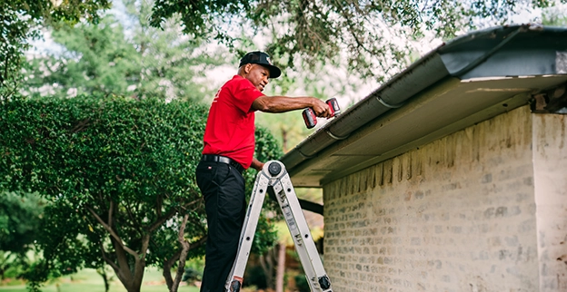 A person in a red shirt stands on a ladder, using a power drill to work.