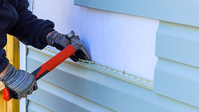 A person wearing gloves is using pliers to adjust siding on a house, focusing on a section of pale blue exterior paneling.