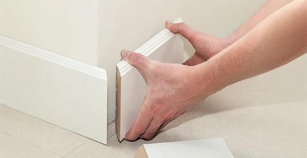 A person installs a trim piece against a wall holding it with both hands near the floor with a section of trim visible nearby.