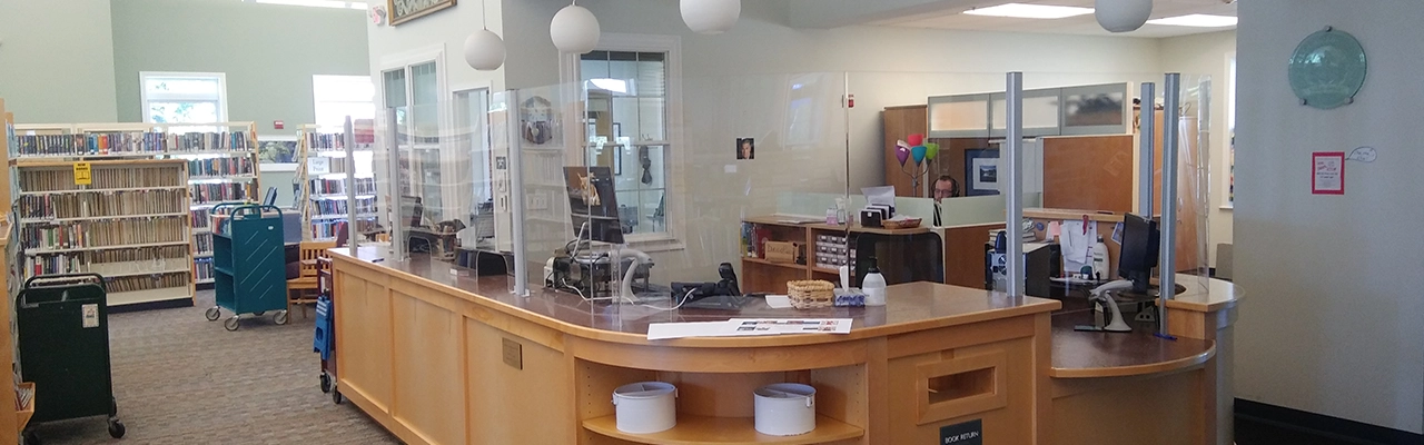 A bright library interior featuring a wooden checkout desk with protective glass.