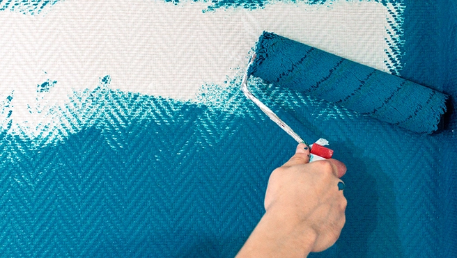 A hand uses a paint roller to apply a vibrant turquoise color to a textured wall.