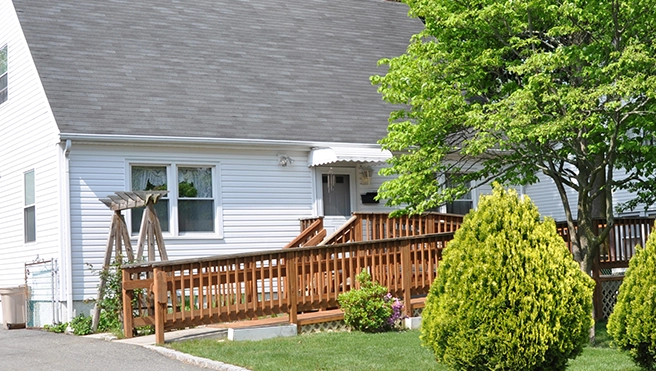 A well-maintained white house with a wooden ramp, surrounded by green shrubs and trees.