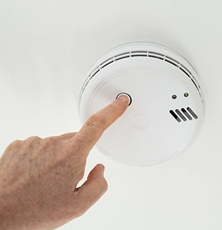 A hand pressing the test button on a smoke detector mounted on a ceiling.