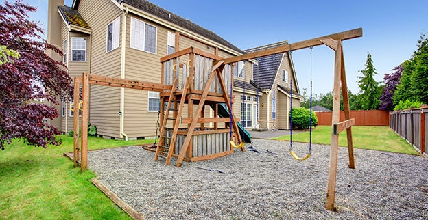 A wooden playground with swings sits on gravel, surrounded by green grass and trees, beside a large beige house.