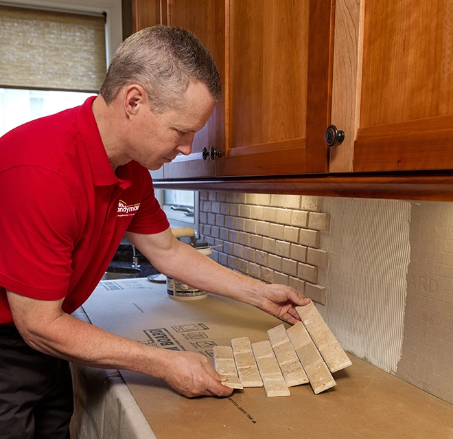 A worker arranges stone tiles on a countertop under wooden cabinets in a kitchen, preparing for installation.