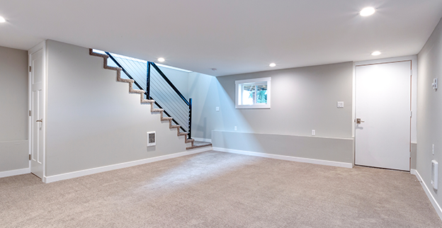 Empty modern basement room with carpeted flooring, and a staircase with a black railing leading up.