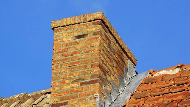 A brick chimney rises from a roof with a mix of gray and orange tiles.