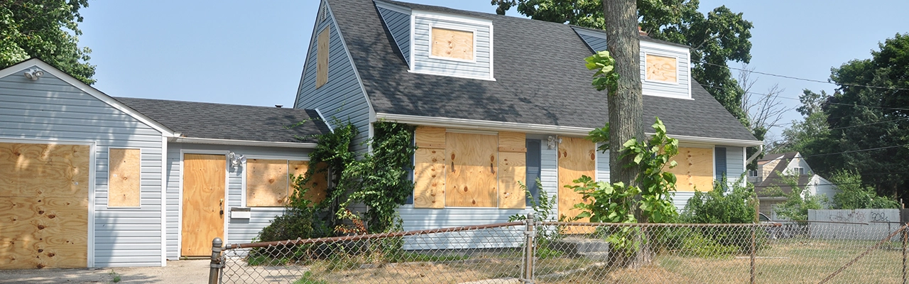 Abandoned house with boarded windows and doors, overgrown greenery and a rusty fence.
