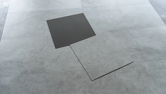 Two square tiles, one dark gray and one light gray, are placed on a light gray textured flooring.