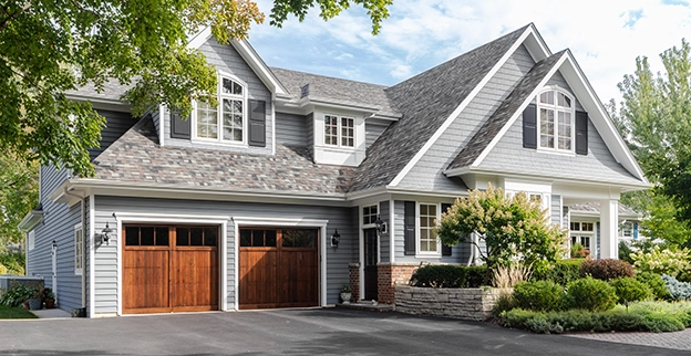A charming gray house with a steep roof, wooden garage doors, and lush landscaping under a sunny sky.