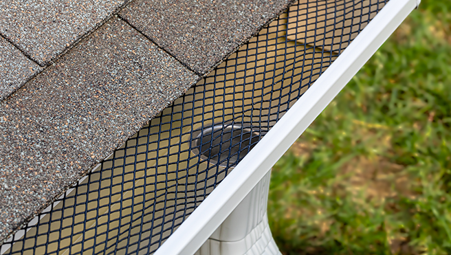 Close-up of a gutter guard installed on a roof, featuring a diamond-patterned mesh over shingles, with a drainage outlet.