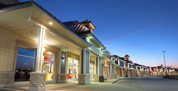 A modern shopping plaza at dusk featuring well-lit storefronts with large windows and a clear sky backdrop.