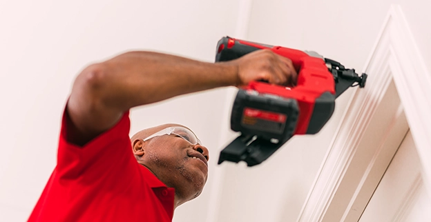 A man in a red shirt and safety goggles uses a power tool to install trim on a wall.