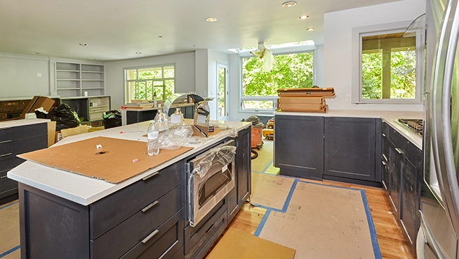 A partially renovated kitchen with dark cabinets, light countertops, construction debris and large windows overlooking greenery.
