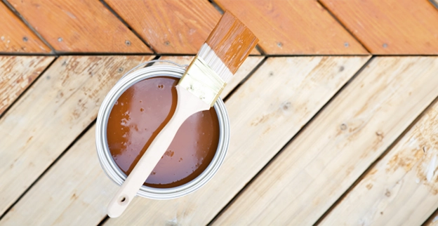 A paint can filled with brown paint and a brush rests on a wooden deck with a herringbone pattern.