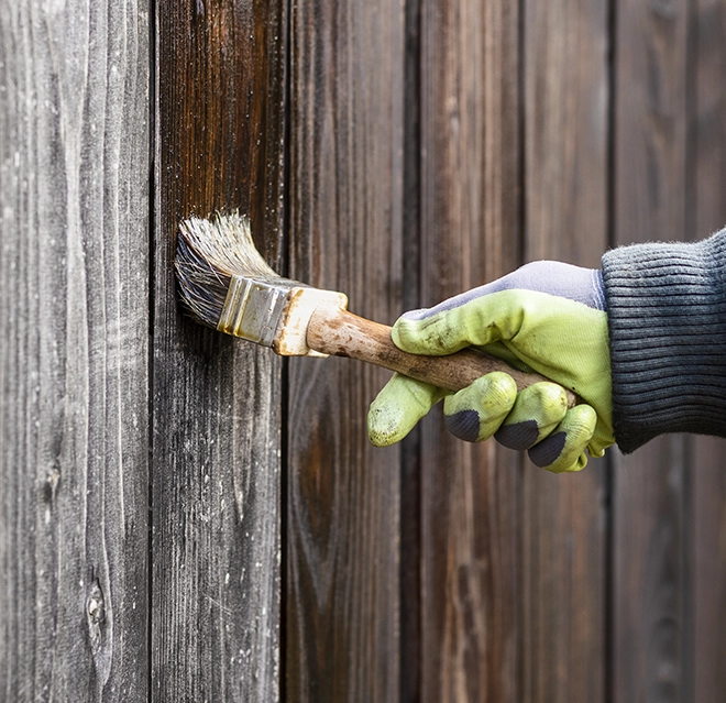 A gloved hand holds a paintbrush, applying a dark finish to a wooden fence panel.