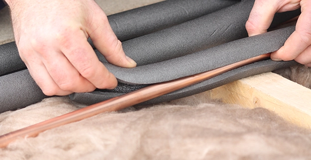 A person’s hands wrap foam insulation around a copper pipe over a soft surface, preparing for installation.