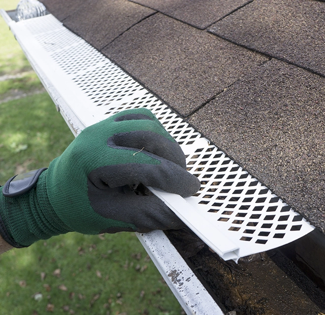 A gloved hand installs a white gutter guard on a roof with brown shingles.
