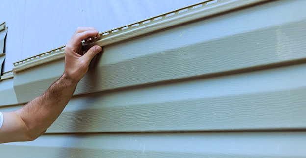A hand is installing paneling on a light green exterior wall, demonstrating home improvement or renovation work.