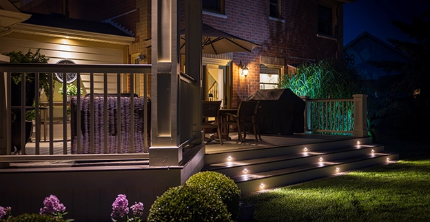 A cozy patio illuminated at night, featuring steps with lights, a grill, and seating, surrounded by greenery and blooming flowers.