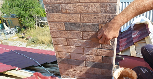 A person applies mortar to a brick-patterned chimney on a rooftop.