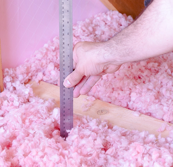 A hand holding a metal ruler measures pink insulation material in an attic space on a wooden beam.