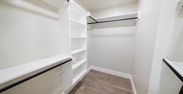 A spacious empty closet featuring white shelving units and black hanging rods against light walls and a wooden floor.
