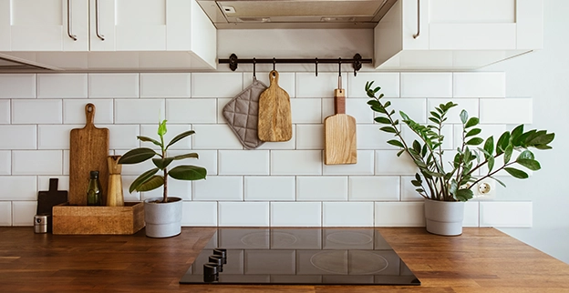 Modern kitchen with wooden countertops, hanging utensils, potted plants, and a sleek cooktop against white subway tile walls.