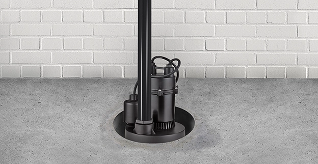 A black sump pump sits in a basin on a concrete floor, near a textured white brick wall.