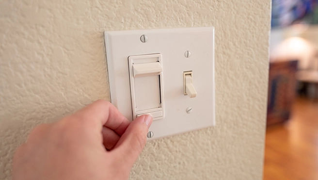 A hand reaches for a light switch on a white wall.
