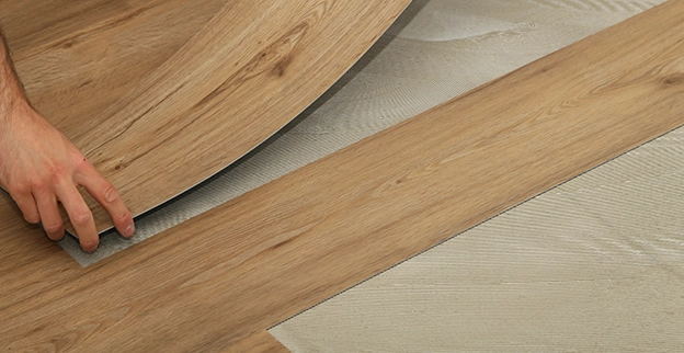 A person installs laminate flooring, carefully aligning a wood plank with the patterned underlayment beneath.