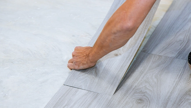 A person's hand carefully aligns a plank of light gray laminate flooring while installing it over a smooth.