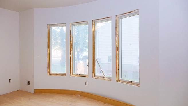Interior of a room featuring four large, unframed windows with a light wood floor and unfinished white walls.