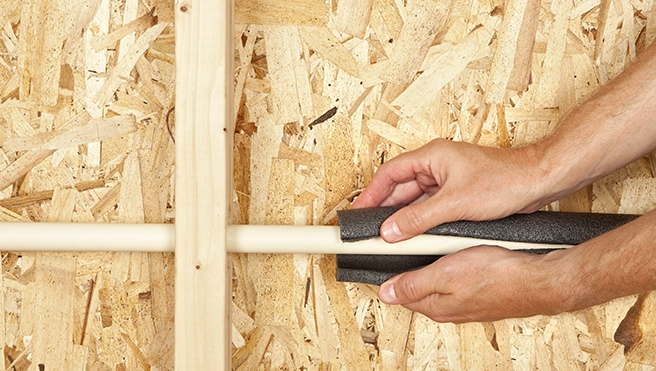 A person wrapping foam insulation around a white plastic pipe mounted on an oriented strand board wall.