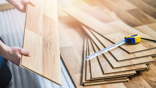 A person holds a wooden flooring plank next to stacked planks and a measuring tape on a partially finished wooden floor.