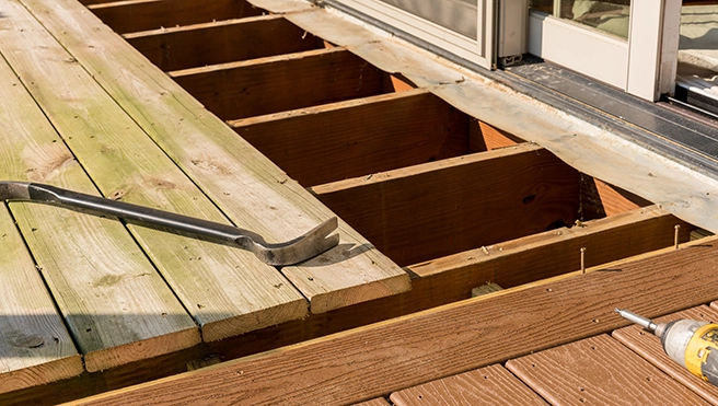 A partially dismantled wooden deck showing exposed beams, a pry bar, and a drill, with a sliding glass door in the background.