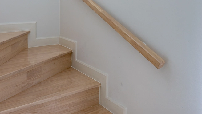 Wooden stairs with smooth, light-colored steps and a natural wood handrail against a plain white wall.
