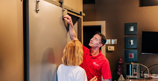 A technician in demonstrates an installation or repair process to a woman in a cozy indoor setting.