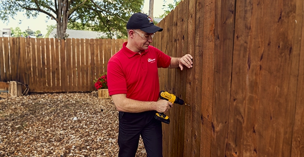 A handyman using a power drill to secure wooden planks against a wooden fence in a garden setting.