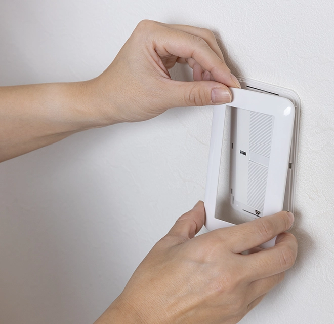 A person installs a white frame around a wall-mounted device.