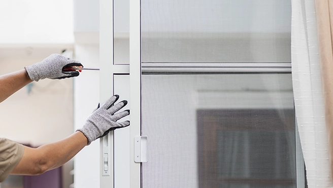 A person in gray gloves adjusts a sliding screen door, using a small tool to ensure proper fitment.