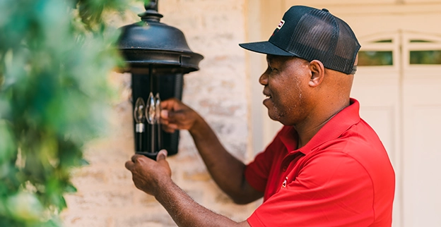A person in a red shirt adjusts an outdoor lantern attached to a stone wall, surrounded by greenery.
