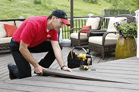 A worker in a red shirt kneels on a deck, using a tool to repair flooring.