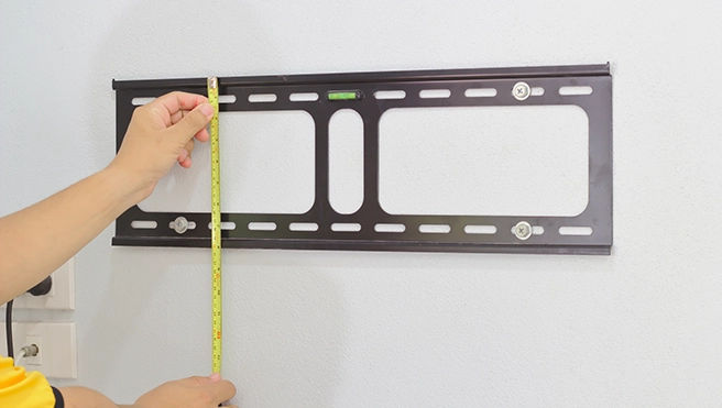 A person measuring the height of a black TV wall mount bracket on a wall with a measuring tape, ensuring proper installation.